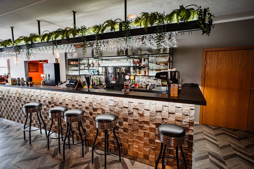 Stylish bar with hanging glassware, leafy plants, wood-textured front panels, and black leather stools on a chevron-patterned floor.
