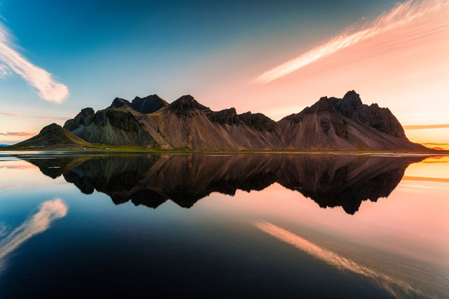 Vestrahorn Mountain reflected in calm water at sunset, with vibrant skies and dramatic peaks in southeastern Iceland.