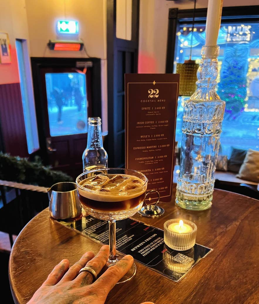 Espresso martini on a round wooden table with a lit candle, cocktail menu, and hand reaching for the glass in a cozy bar setting.