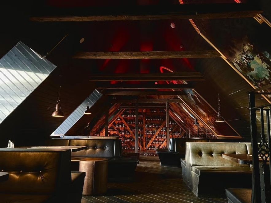 Schummrig beleuchtete Loft-Bar mit Ledersesseln, freiliegenden Holzbalken, schr&auml;gem Dach und einer R&uuml;ckwand voller Regale mit Spirituosenflaschen