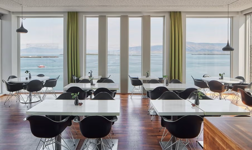 Modern dining area with black chairs, white tables, and large windows overlooking a calm sea and distant mountains.
