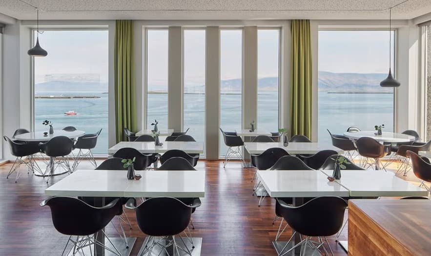 Moderne eetruimte met zwarte stoelen, witte tafels en grote ramen met uitzicht op een kalme zee en verre bergen.