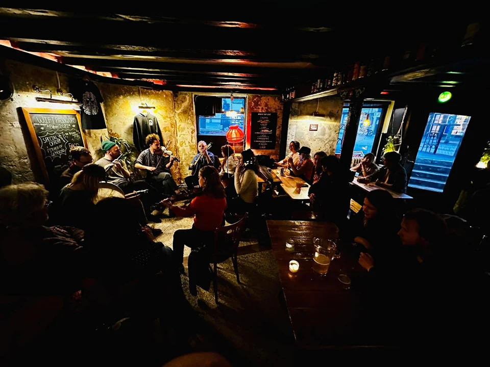 Cozy pub with dim lighting, a live folk band playing fiddles and flutes, and patrons gathered closely around wooden tables.