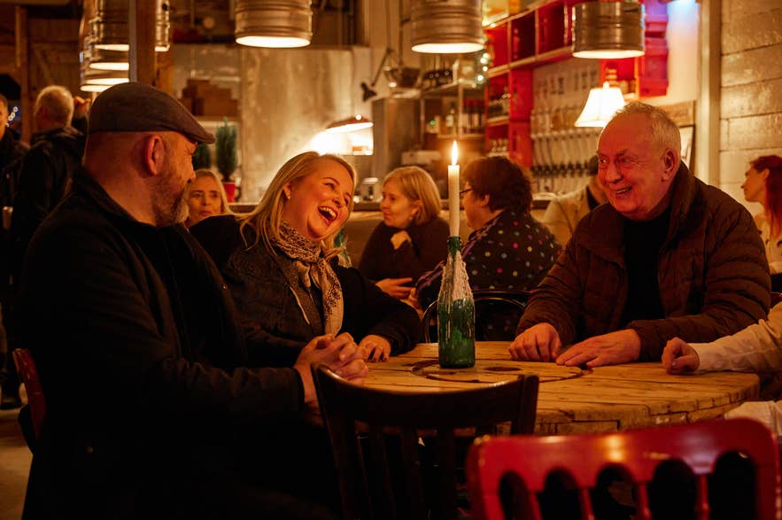Group of people laughing and chatting around a candlelit wooden table in a cozy, warmly lit taproom.