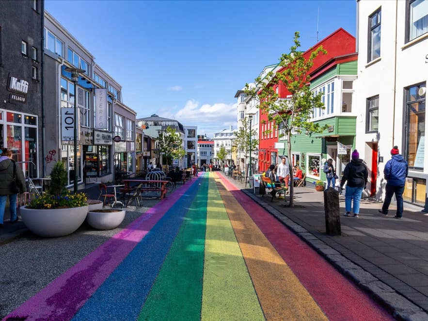 Colorful rainbow-painted street in downtown Reykjavik with people walking past shops, cafes, and buildings.