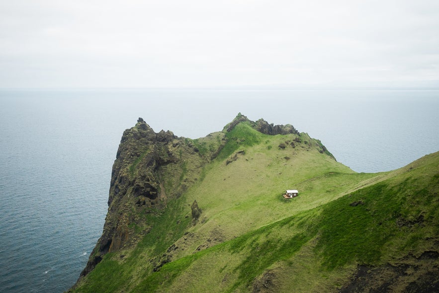 A grassy peak of Blatindur mountain on Heimaey island with a small hut and steep cliffs overlooking the ocean.