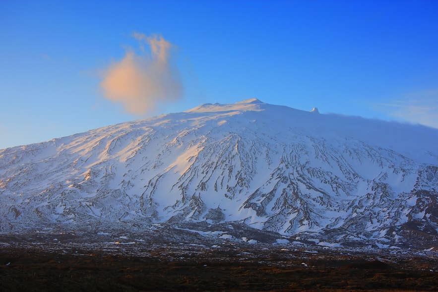 澄み切った青空の下、柔らかな日差しを浴びて雪に覆われたスナイフェルスヨークトル火山。山頂の上には、一筋の雲が漂っている。
