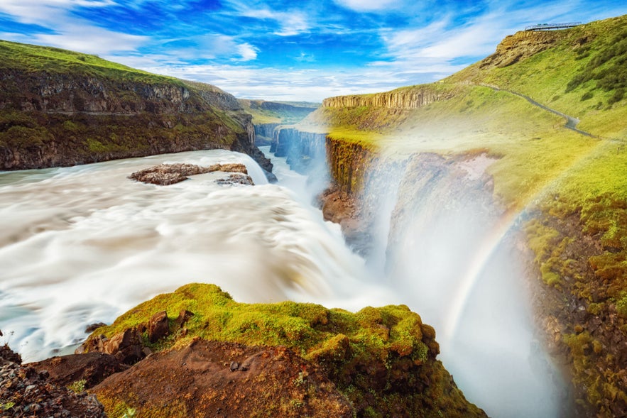 Powerful Gullfoss waterfall plunges in two stages into a canyon, with mist and a rainbow under a bright blue Icelandic sky.