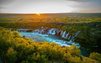Sunset over Hraunfossar Waterfall with turquoise water flowing through lava rock and golden autumn trees in West Iceland.