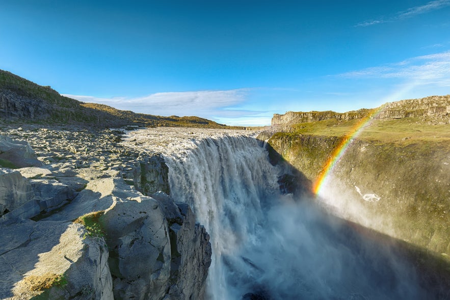 Dettifoss Waterfall thunders into a rocky canyon, creating mist and a vivid rainbow under a clear blue sky in North Iceland.