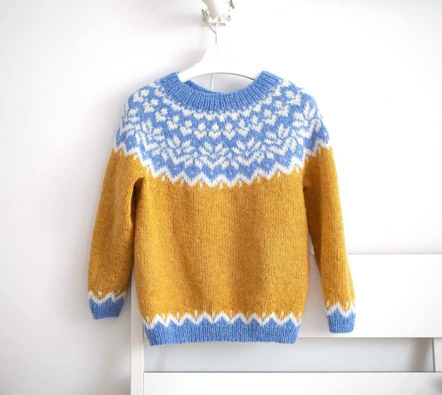 Ein senfgelber und blauer Lopapeysa-Pullover aus Island mit traditionellem Muster hängt an einer weißen Wandhalterung.