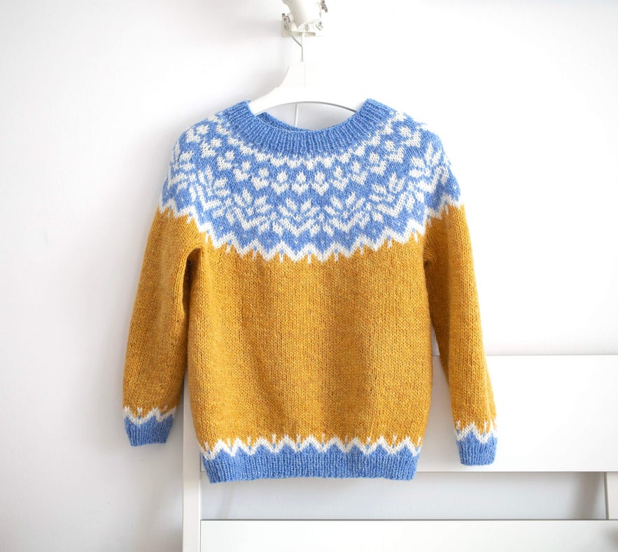 Mustard yellow and blue Icelandic lopapeysa sweater with traditional pattern, hanging on a white wall-mounted hanger.