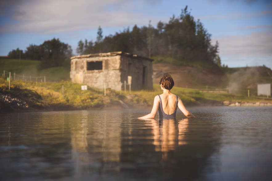 Persoon baadt in de Secret Lagoon (Gamla Laugin) in Fludir, Zuid-IJsland, met geothermische stoom en een historisch stenen gebouw.
