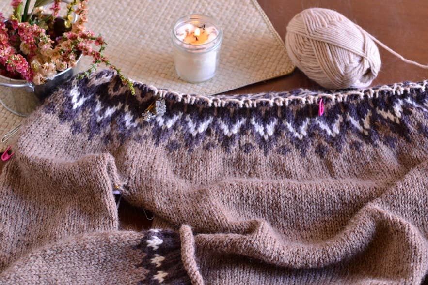 Ein handgestrickter Lopapeysa-Pullover in Arbeit mit klassischen isländischen Mustern, umgeben von Garn, Stricknadeln und Kerzenlicht.