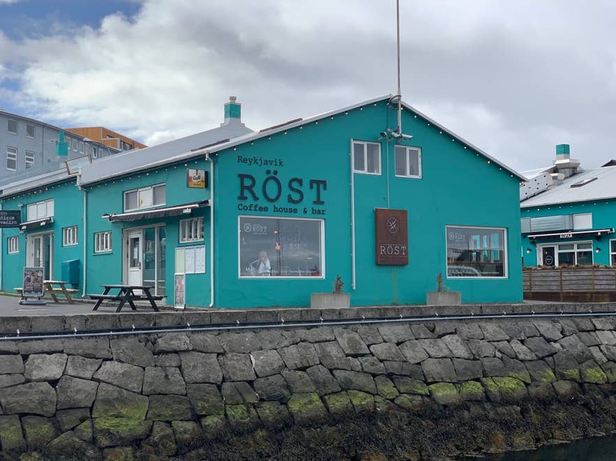 The turquoise Rost Coffee House & Bar in Reykjavik sits along the harborfront, with outdoor seating and stone quay in view.