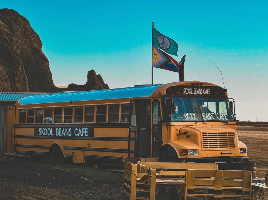 Skool Beans Cafe à Vik, en Islande, est dans un ancien bus scolaire jaune reconverti, avec des drapeaux flottant devant un paysage montagneux.