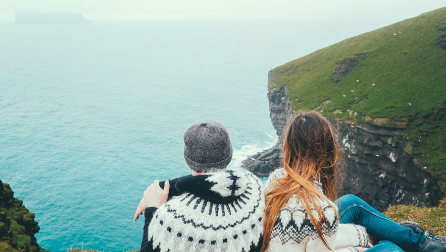 Couple wearing traditional lopapeysa sweaters sits on Icelandic cliffside, overlooking ocean and misty coastal landscape.