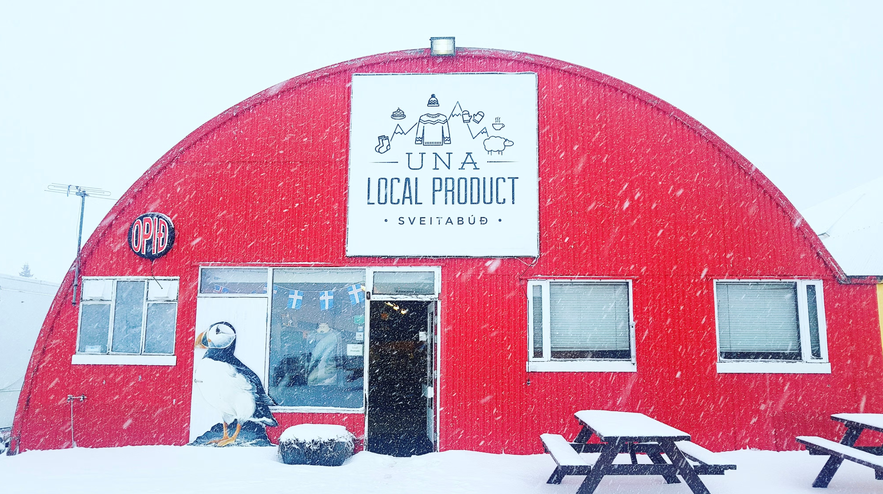 Snow falls outside UNA Local Product, a red arched gift shop with a puffin display and picnic tables covered in snow.