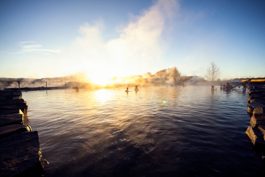Visitors are bathing in the Secret Lagoon in Iceland under a clear blue sky with the setting sun in the background.