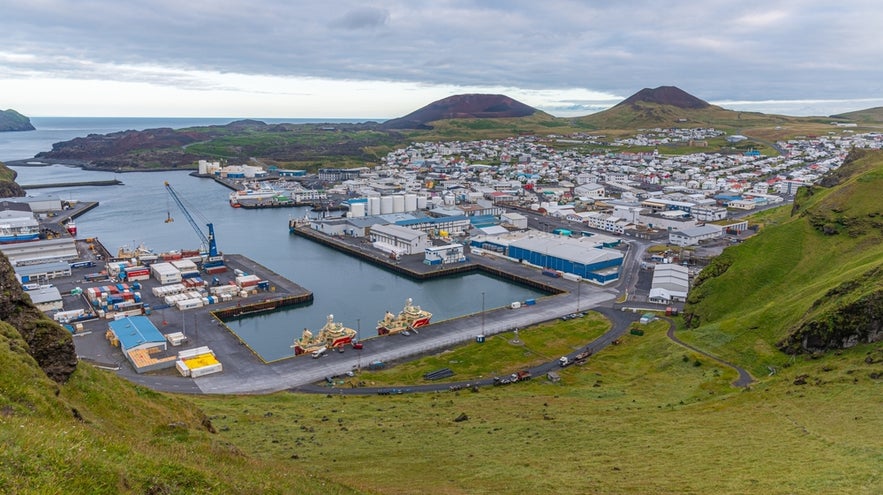Heimaey town and harbor in the Westman Islands, Iceland, with volcanic hills and fishing boats under cloudy skies.