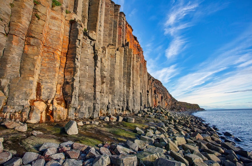 Basalt columns at Kalfshamarsvik, Iceland, rising along the rocky shoreline under a blue sky with wispy clouds.