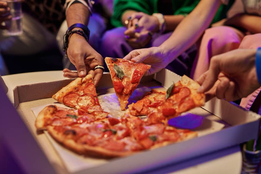 Close-up of slices of appetizing pizza in square cardboard box and hands of young friends taking them