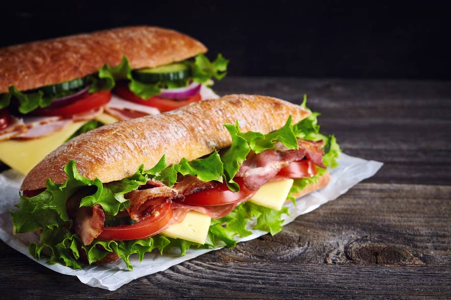 Two fresh submarine sandwiches with ham, cheese, bacon, tomatoes, lettuce, cucumbers and onions on dark wooden background