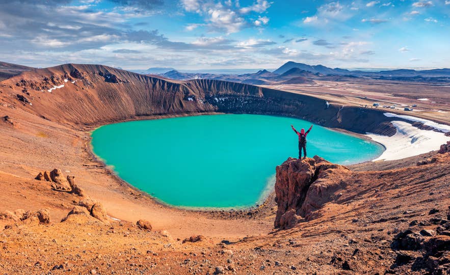 Hiker stands by the vivid turquoise Viti Crater Lake in Iceland’s Krafla volcanic area under a partly cloudy sky.