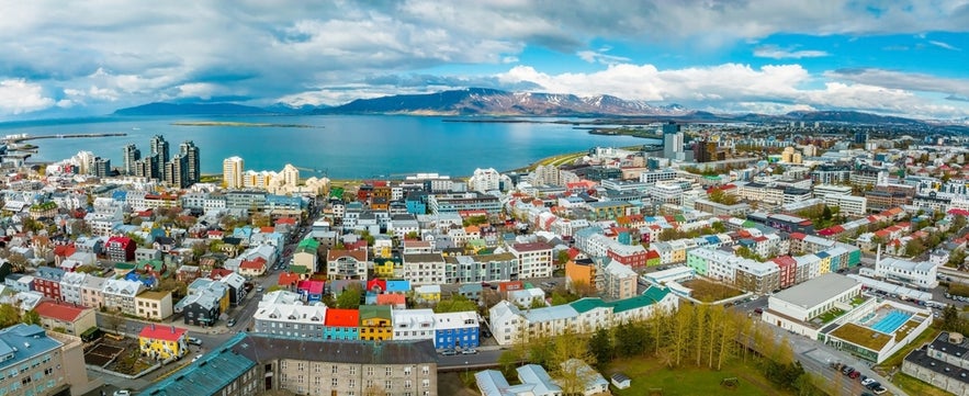 Aerial view of Reykjavik, showing colorful buildings, coastline, and distant mountains under a partly cloudy sky.