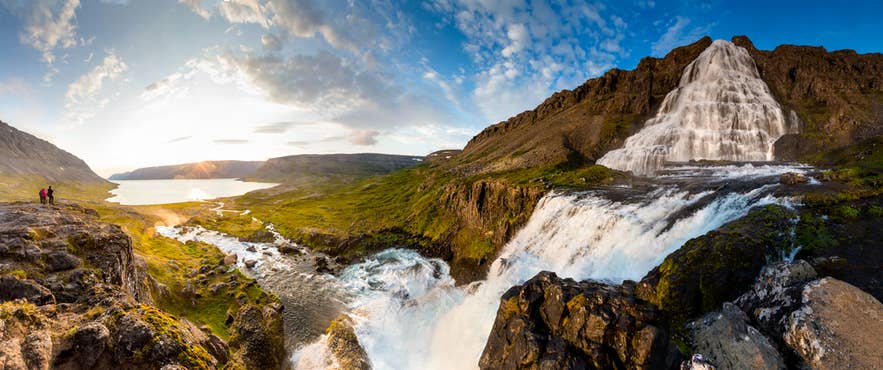 Panoramic view of Dynjandi Waterfall in Iceland cascading down cliffs into a river, with hikers and fjord scenery at sunset.