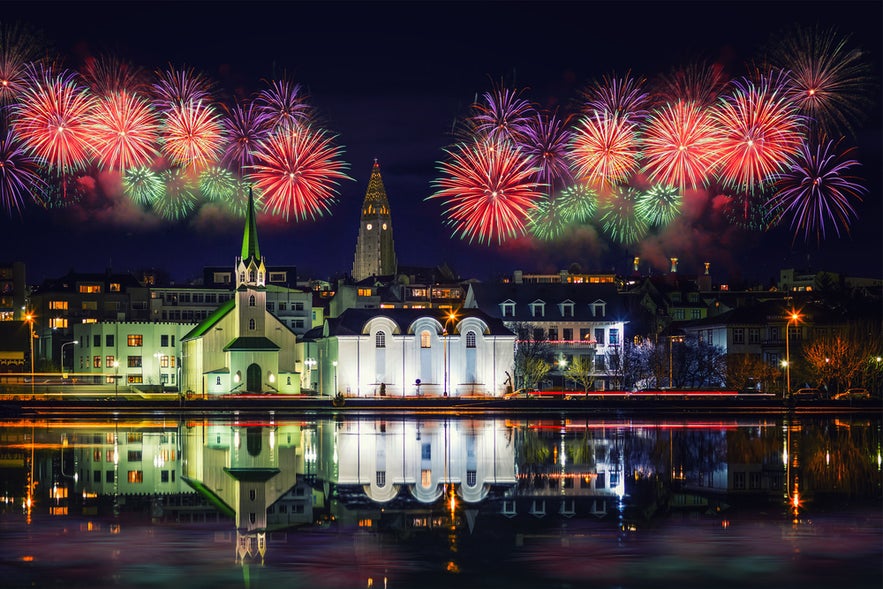 Colorful fireworks light up the night sky over Reykjavik, reflecting in the water with city buildings in the foreground.