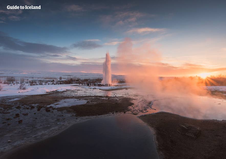 Strokkur geyser erupting at sunset in Iceland’s Golden Circle with steam, snow, and tourists watching nearby.