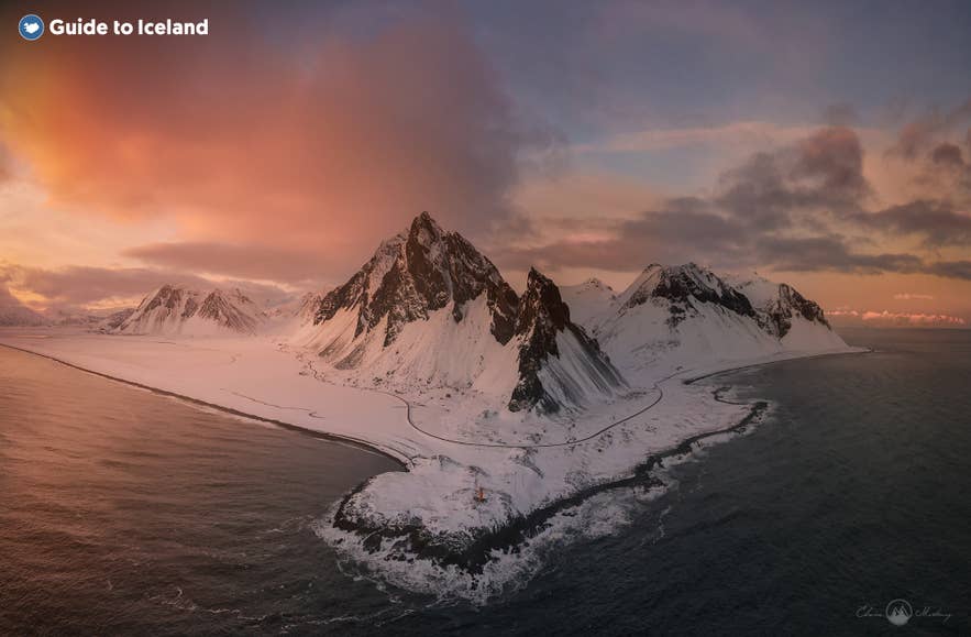 Snötäckta Vestrahorn i solnedgång på Island, med dramatiska moln och havsvågor längs stranden