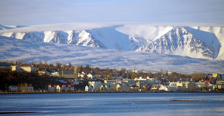 Snowy mountains behind the town of Akureyri, with Akureyrarkirkja church and colorful buildings along the calm waterfront.
