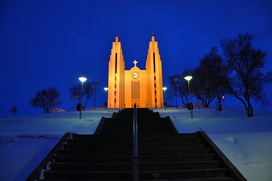 Akureyrarkirkja church in Akureyri glowing warmly at night, seen from snowy steps under a deep blue winter sky.