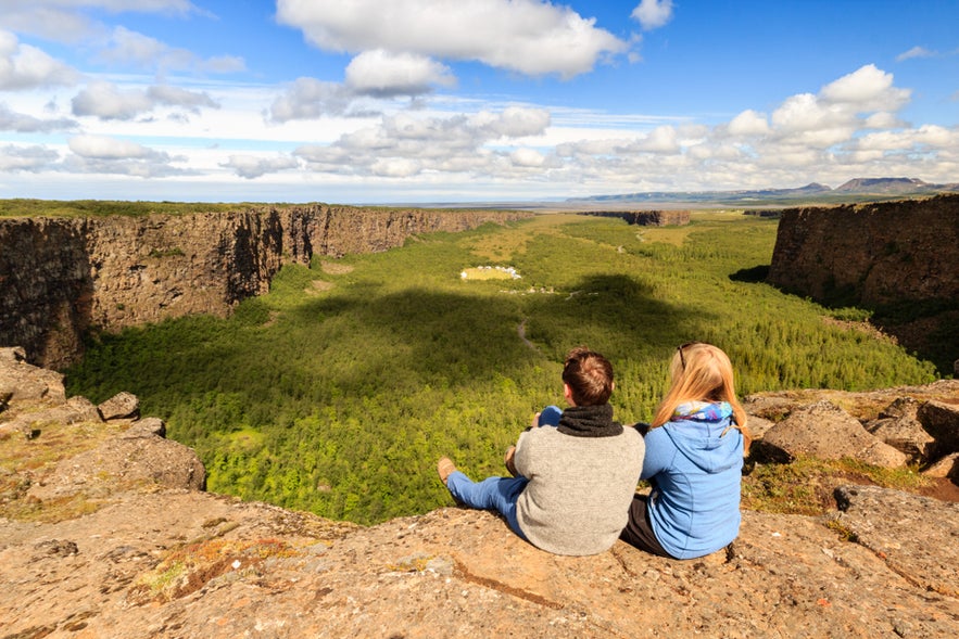 Two hikers sit atop Asbyrgi Canyon in North Iceland, enjoying the scenic view of the horseshoe-shaped canyon below.