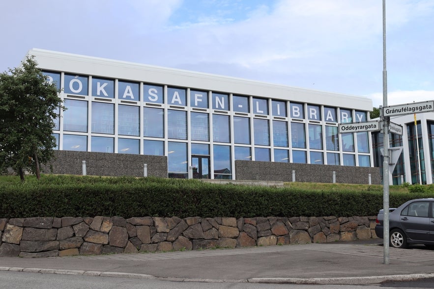Modern glass-fronted building of Akureyri Municipal Library with street signs and parked car in foreground.