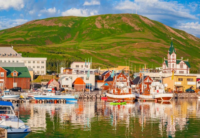 Colorful harbor with boats, traditional houses, and a green hill in the background in Husavik, Iceland.