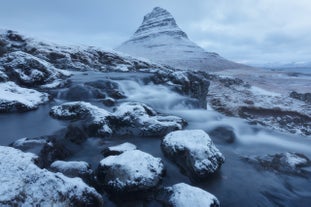 Kirkjufell mountain shaped like a steep cone beside Kirkjufellsfoss waterfall, with snow-covered ground and partially frozen cascades.