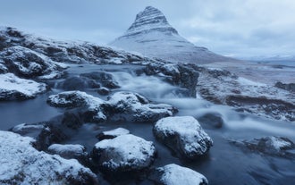 Kirkjufell mountain shaped like a steep cone beside Kirkjufellsfoss waterfall, with snow-covered ground and partially frozen cascades.