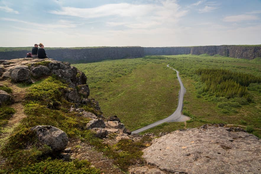 นักท่องเที่ยวสองคนนั่งอยู่บนหน้าผาที่ Asbyrgi Canyon มองถนนที่อยู่ด้านล่างของกำแพงหิน