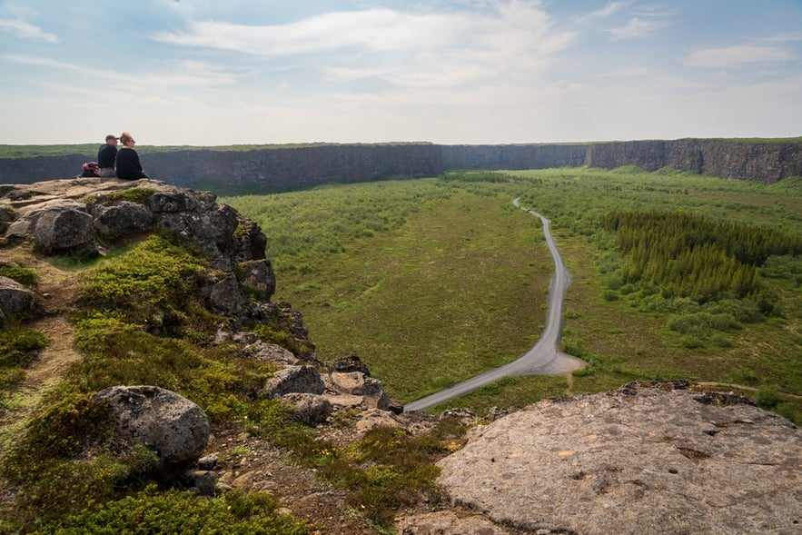 Two visitors sit atop a cliff at Asbyrgi Canyon, overlooking a road below its rocky walls.