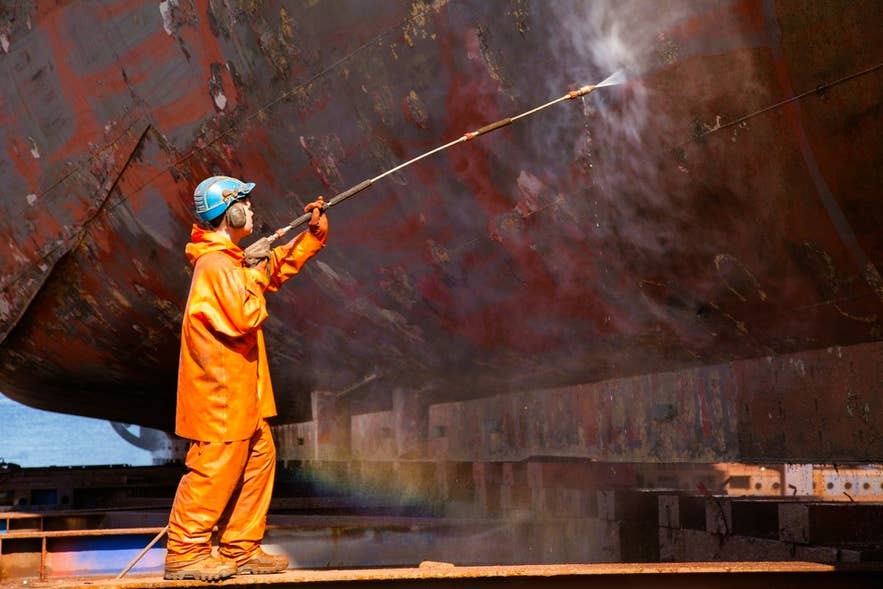 Icelander in orange gear power washes a ship hull, reflecting the nation’s strong work ethic and maritime heritage.