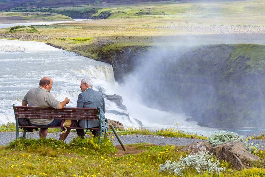 Two Icelandic men in conversation on a bench overlooking a waterfall.