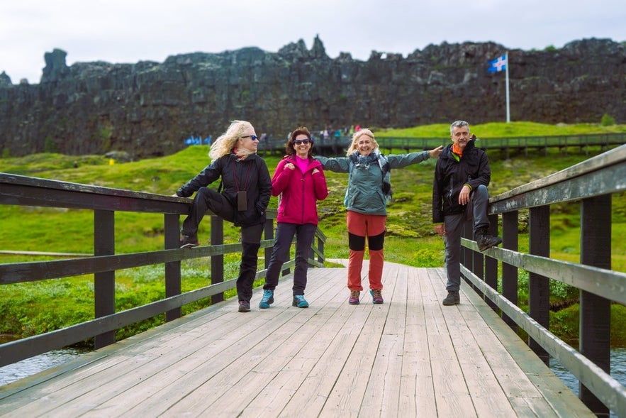 Four Icelanders stand on a wooden bridge at Thingvellir National Park, smiling with the park’s rocky cliffs and Icelandic flag in the background.