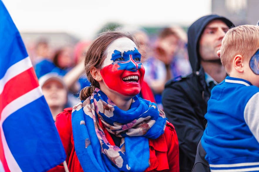An Icelander with the national flag painted on her face smiles brightly in a crowd, holding an Icelandic flag during a public event.