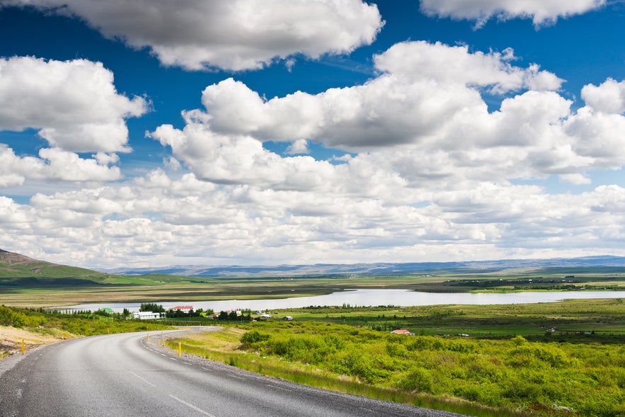 Summer view of Icelandic countryside near Laugarvatn lake, with green fields, a curving road, and bright puffy clouds.