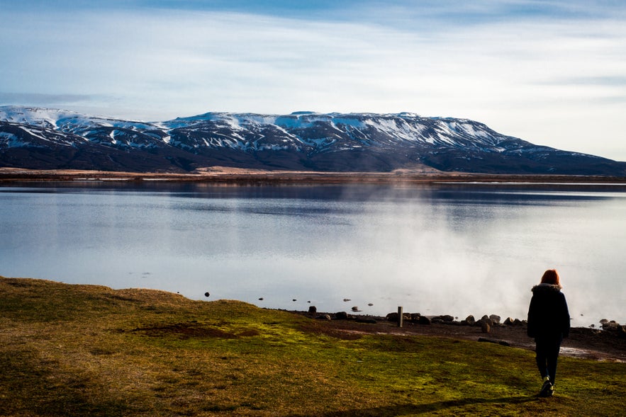 Woman walking by Laugarvatn lake in Iceland, with steam rising near the shore and snow-covered mountains in the background.