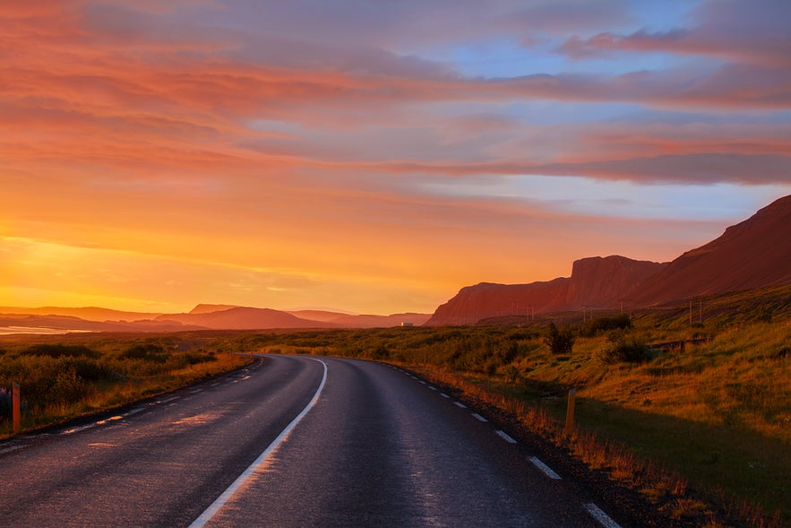 Scenic road in Iceland at sunset, with dramatic orange skies, distant mountains, and lush green landscape along the roadside.