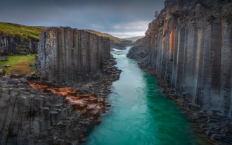 Turquoise Jokulsa a Dal river flowing between towering basalt columns in Studlagil Canyon, East Iceland, under a cloudy sky.